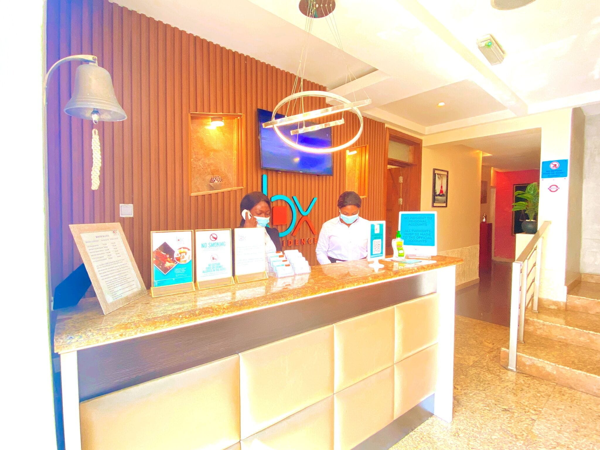 A hotel reception desk with a wooden panel backdrop, a mounted television, and a modern light fixture above.