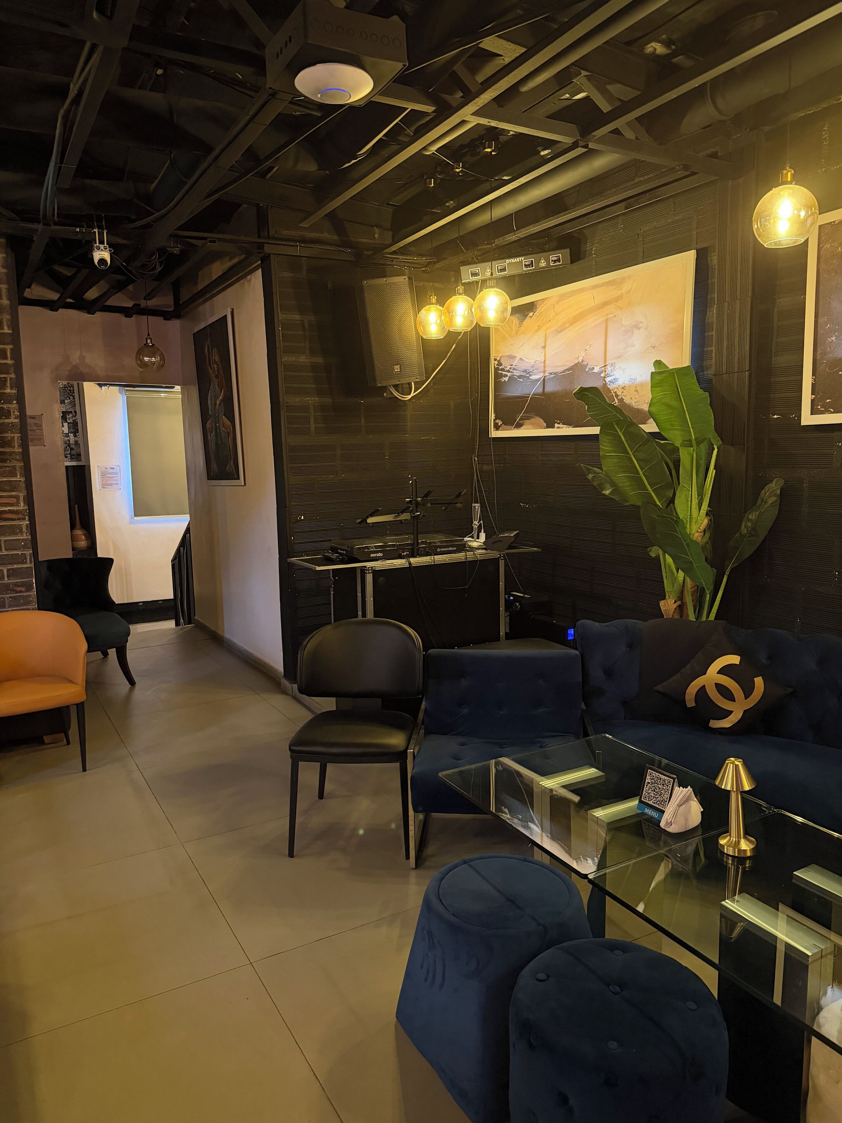 Un espace de détente moderne avec un canapé bleu foncé, une table basse en verre et un fauteuil noir. On y trouve également une grande plante en pot et un haut-parleur fixé au mur.