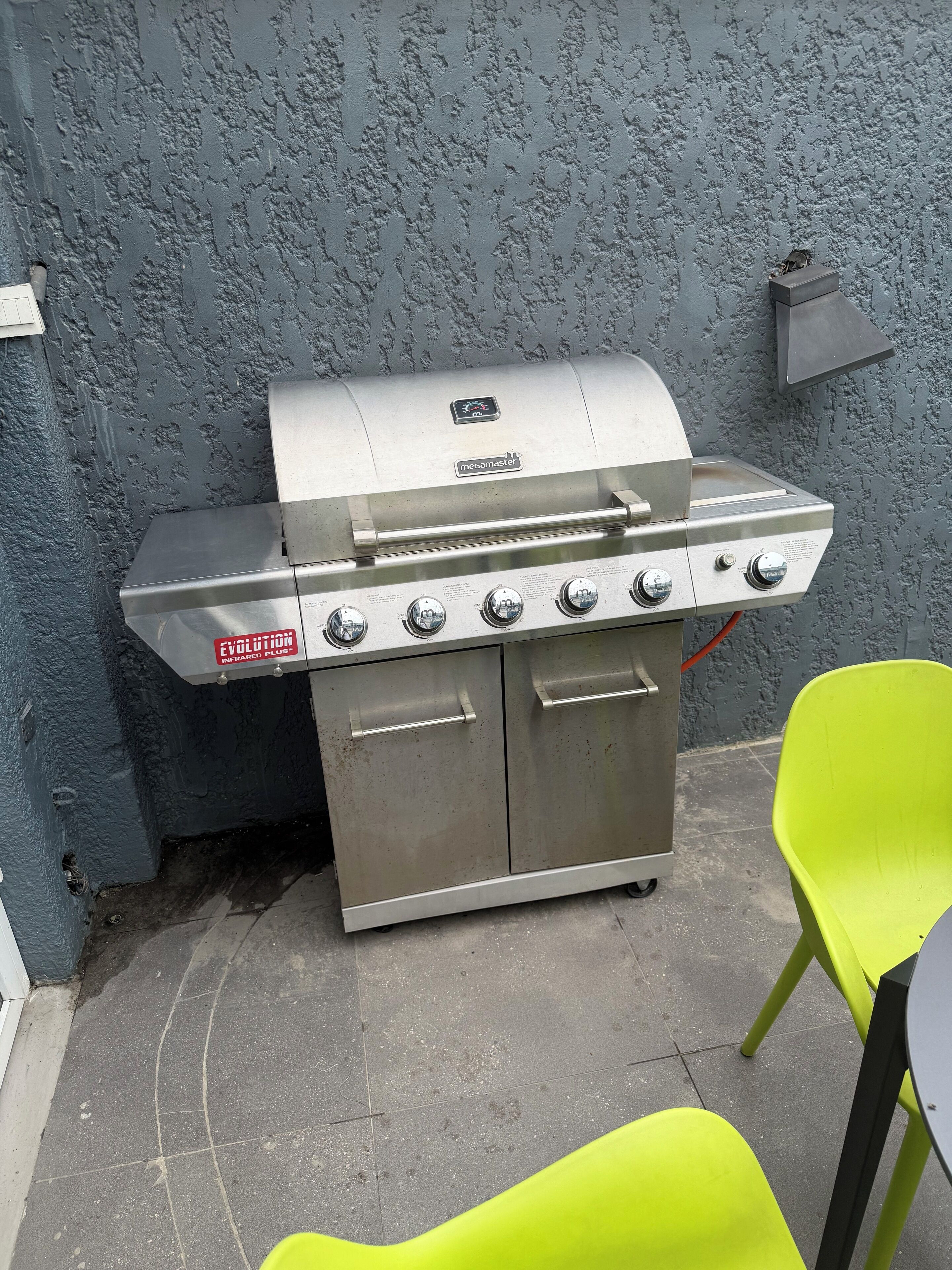 A stainless steel barbecue grill with four burners and a side shelf, positioned against a textured wall.
