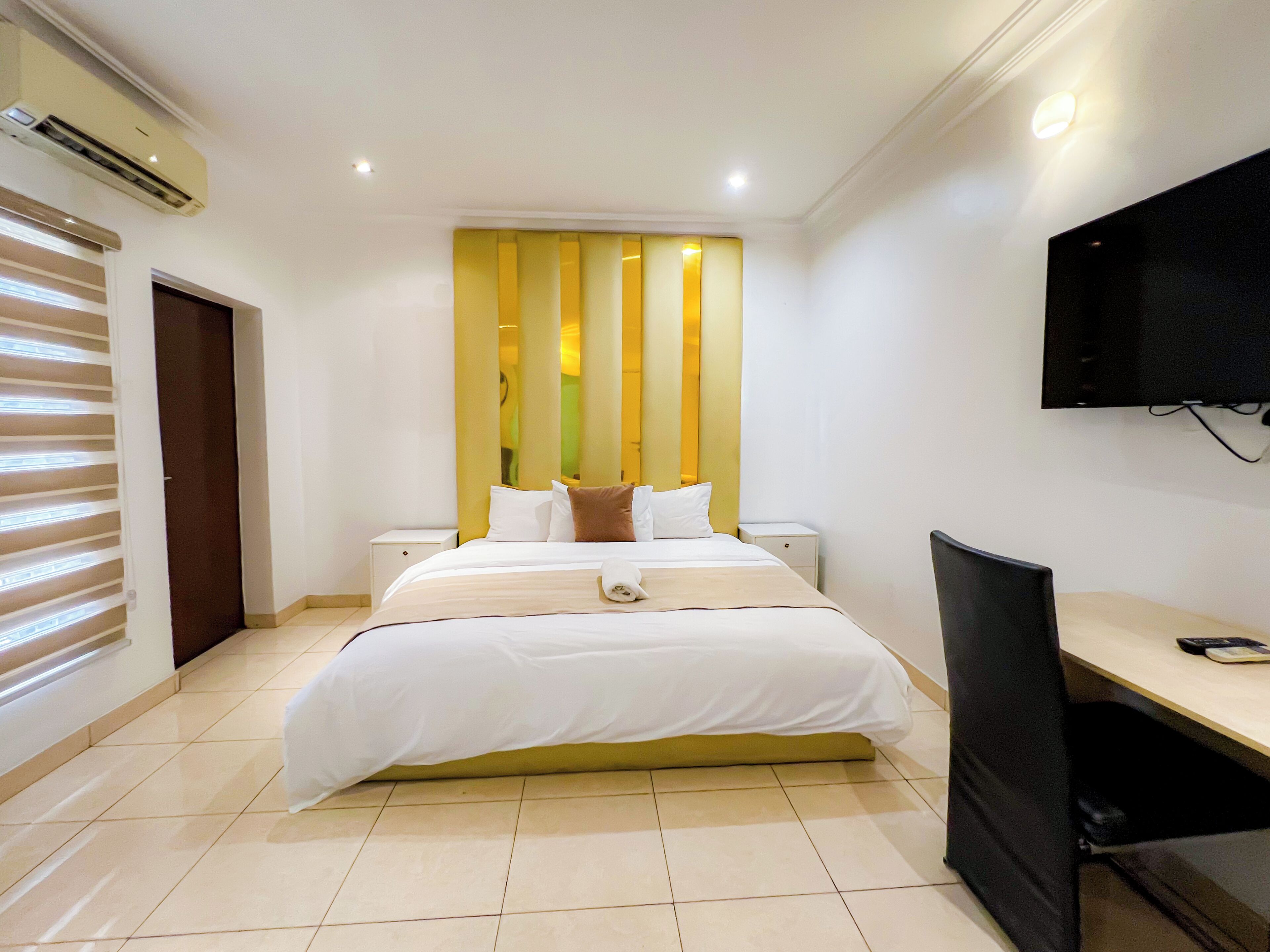 A hotel room with a large bed, a desk, a chair, a TV, and an air conditioning unit.