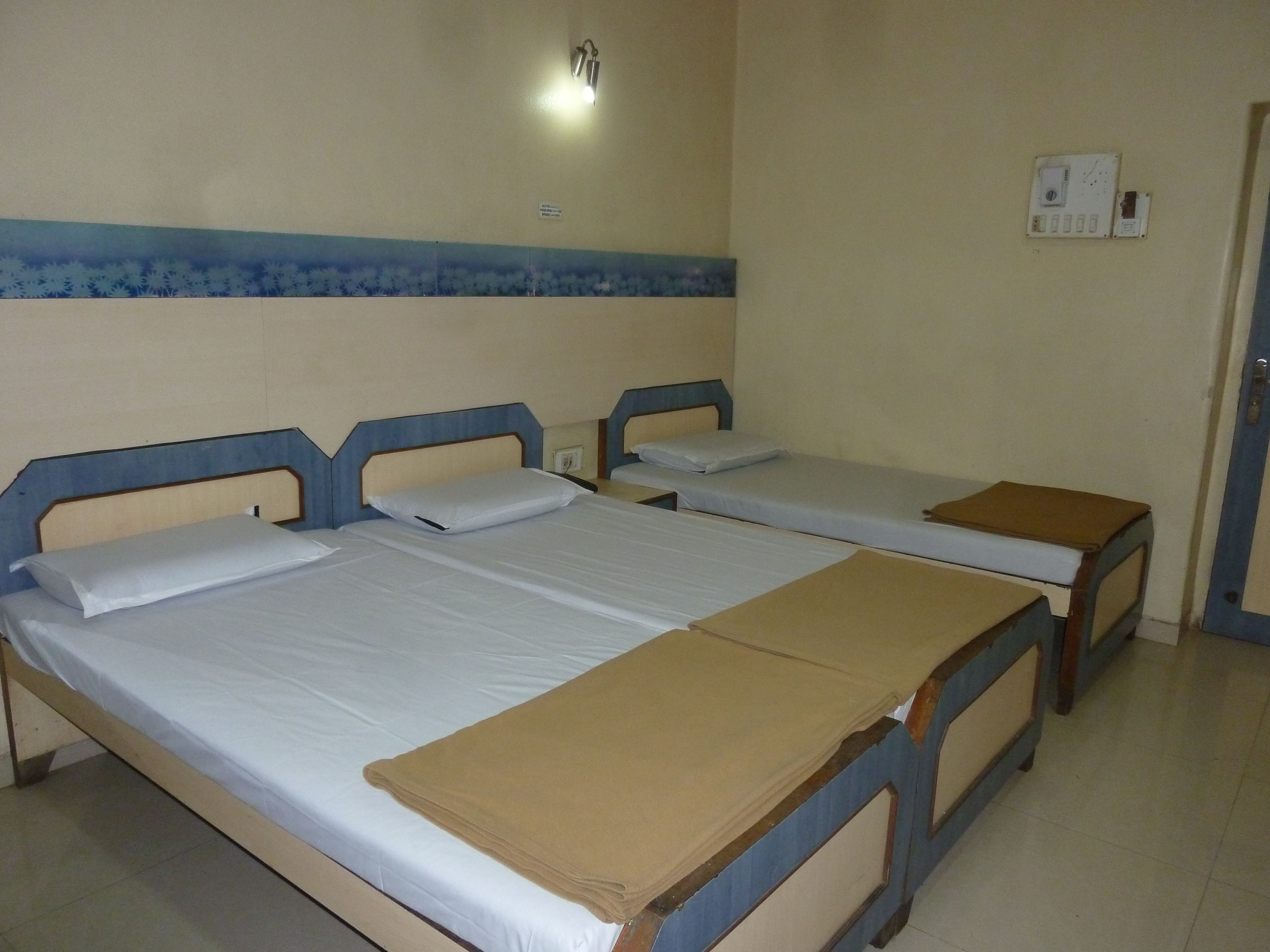 A room with three single beds, each with a wooden frame and white bedding. There is a wall-mounted light fixture above the beds and a thermostat on the wall.