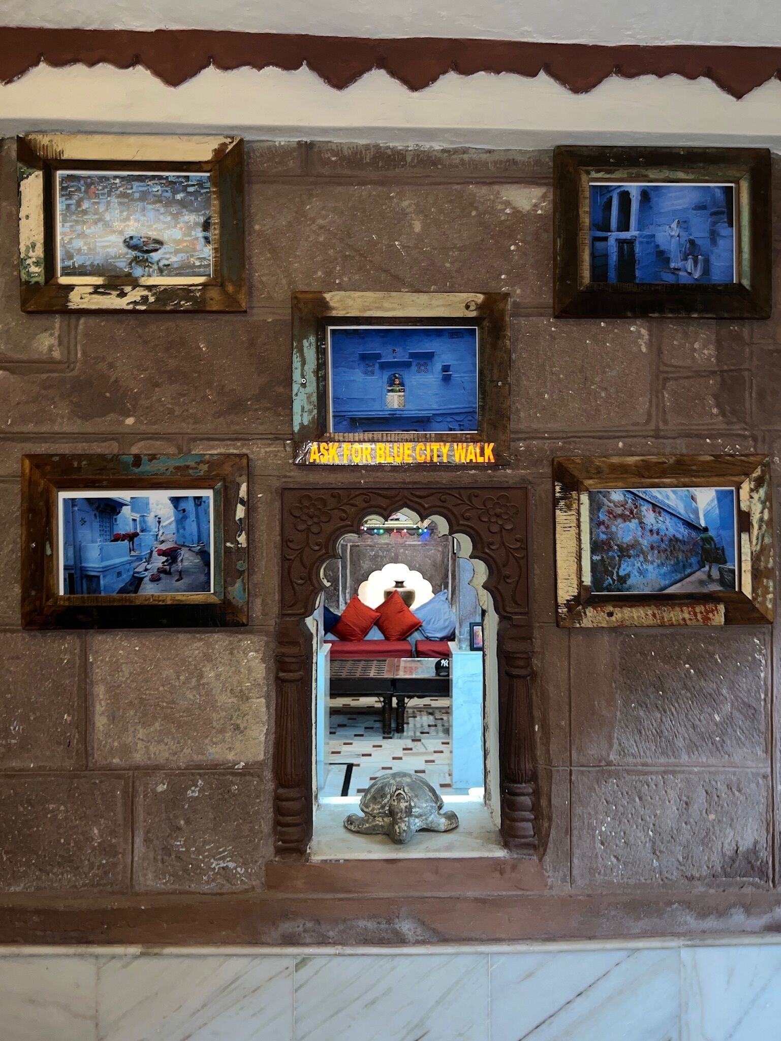 A wall with multiple framed artworks and a central doorway leading to a room with a bed and a statue.