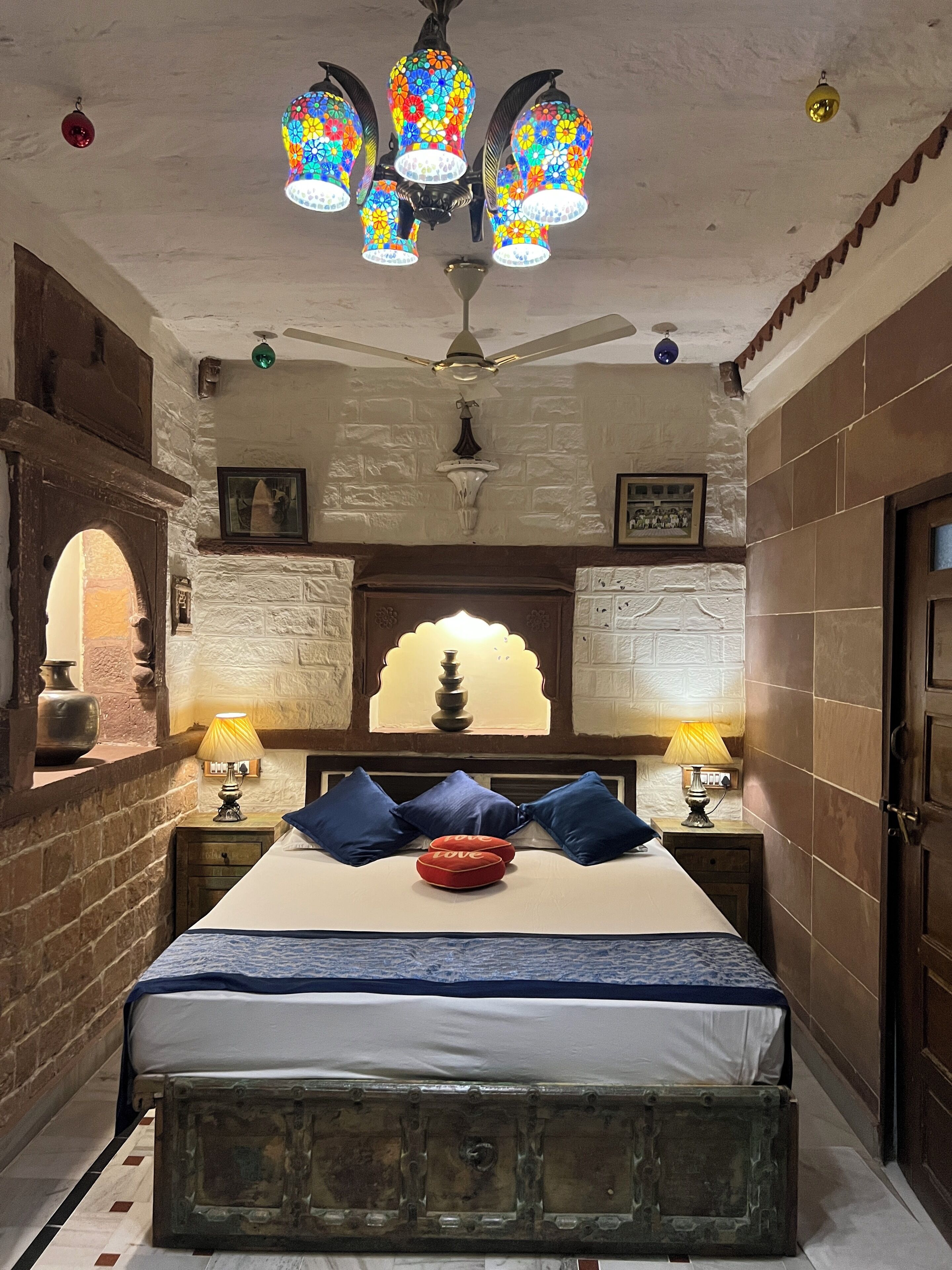 A bedroom with a bed, two bedside lamps, a ceiling fan, and a decorative wall with a window.