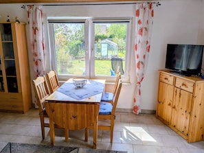 Un área de comedor con una mesa, sillas, un televisor sobre un mueble de madera y una ventana con cortinas.