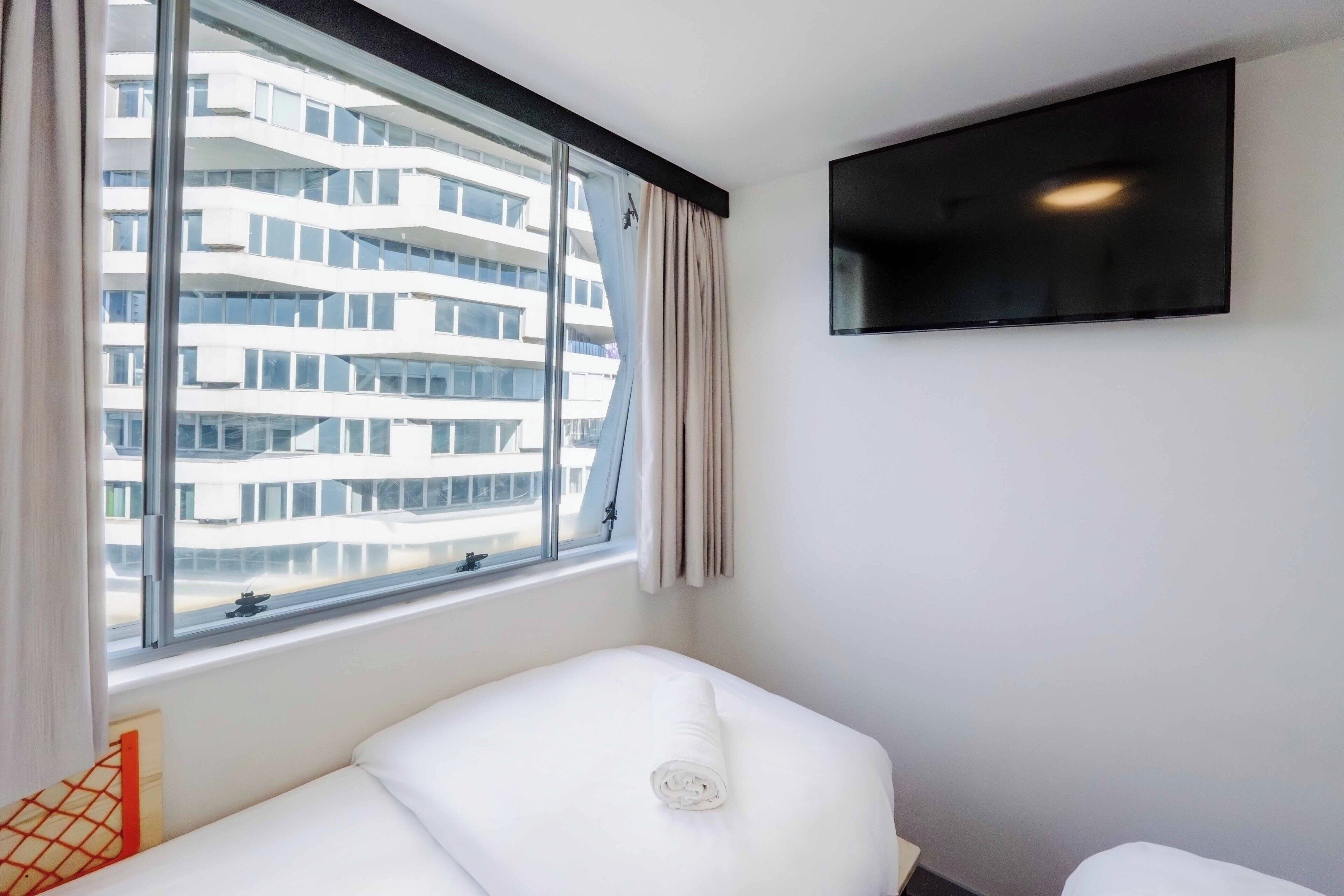 Une chambre d’hôtel dotée d’une grande fenêtre, d’un lit, d’un téléviseur à écran plat fixé au mur et offrant une vue sur un immeuble moderne.
