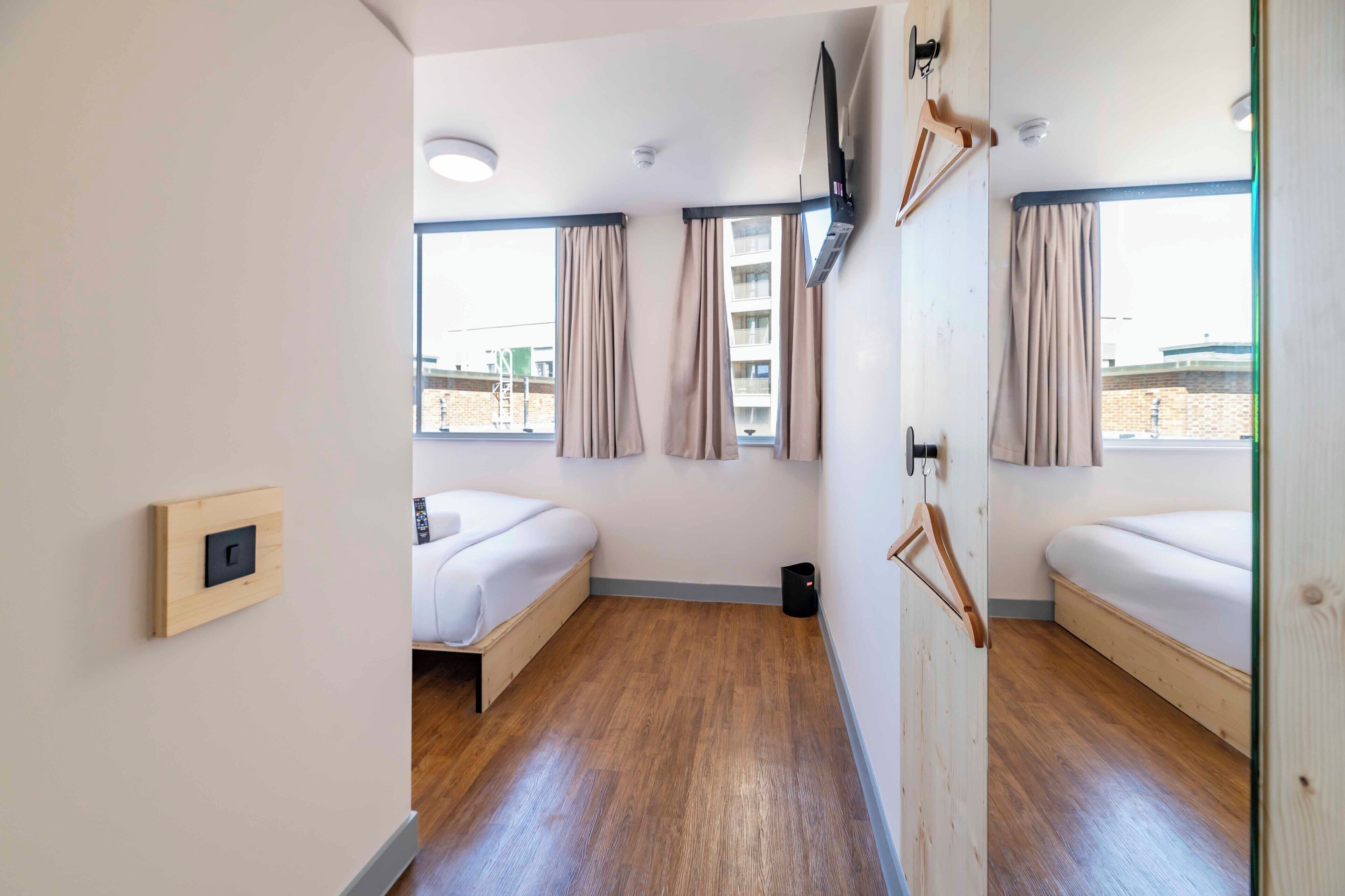 A modern hotel room with two beds, a wooden floor, a flat-screen TV, and large windows with curtains.
