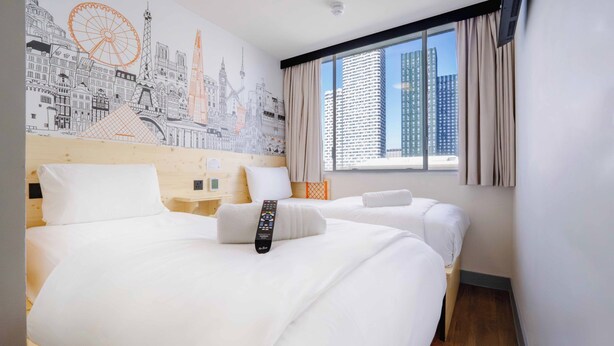 Een hotelkamer met twee bedden, een stadsgezicht muurschildering op de wand, een groot raam met gordijnen en een afstandsbediening op het bed.