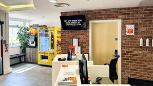 Een receptiezone met een bakstenen muur, een computerbureau, een automaat en een bord met een korting van 10% voor directe boekingen.