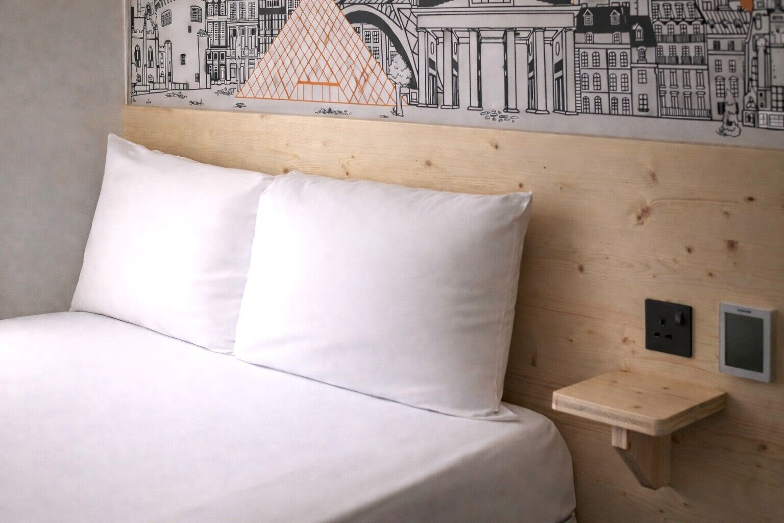 Ein Hotelzimmer mit einem hölzernen Kopfteil, einem Bett mit weißer Bettwäsche, einem kleinen Nachttisch und einem Wandgemälde, das Stadtgebäude zeigt.