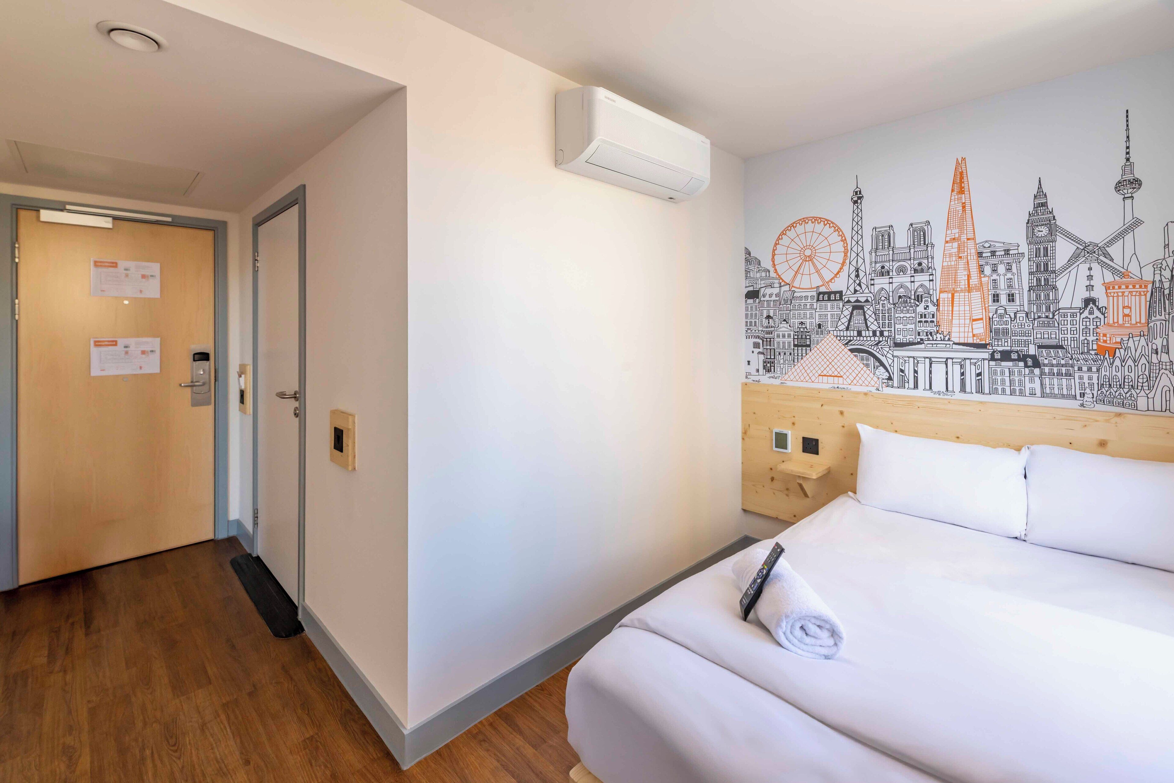 A hotel room with a bed, a cityscape mural, a wooden headboard, and a door with informational cards.