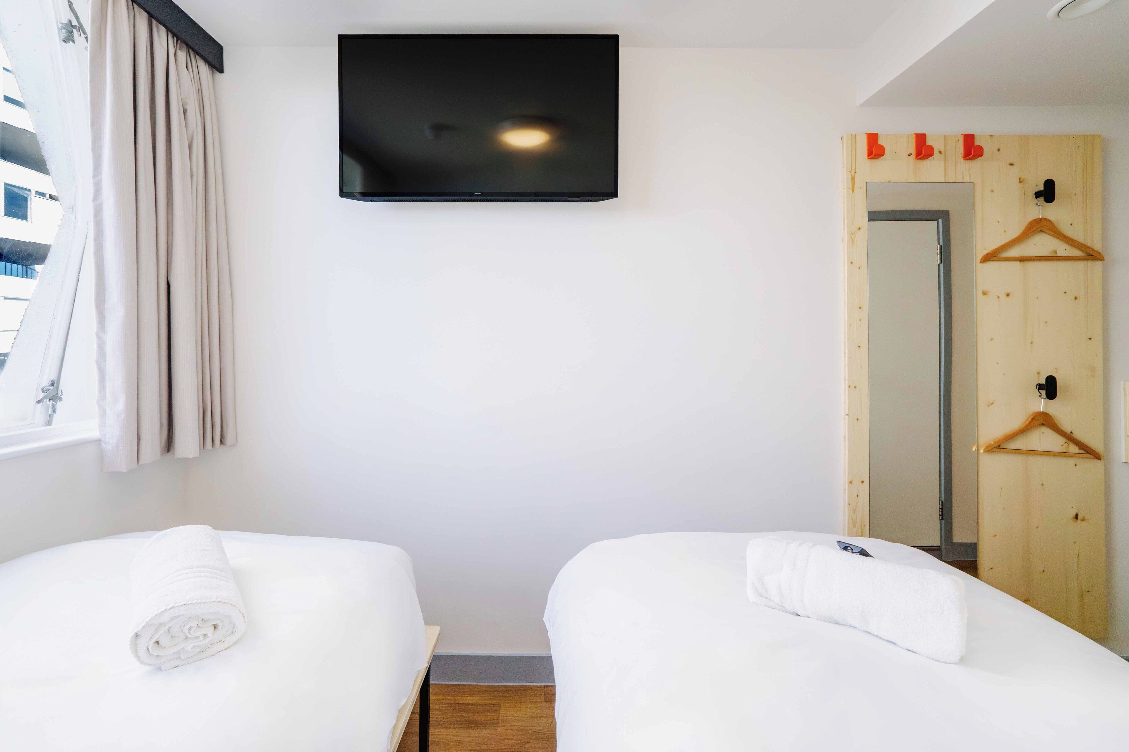 A hotel room with two beds, a TV mounted on the wall, a wooden door with a mirror, and a window with curtains.