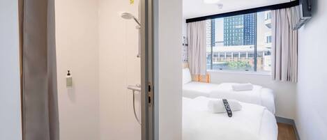Habitación de hotel con ducha, dos camas, ventana con vista a edificios y televisor montado.