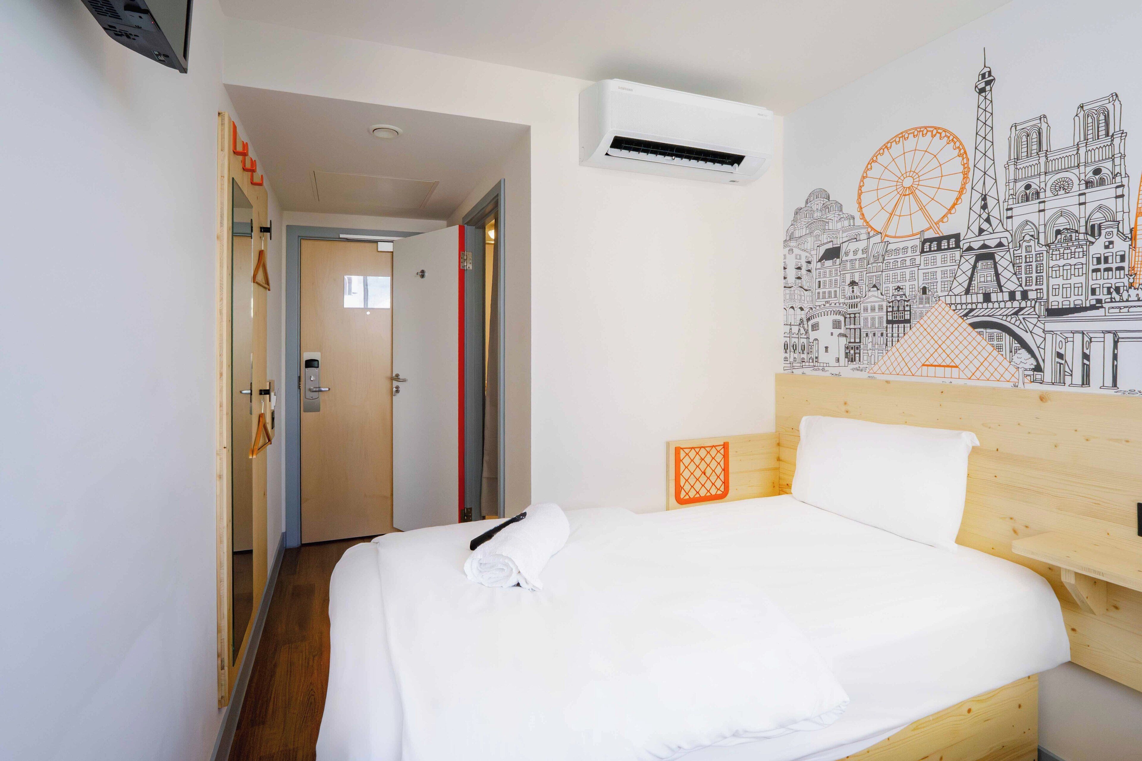 A hotel room with a neatly made bed, an air conditioning unit, a wooden headboard, and a cityscape mural on the wall.