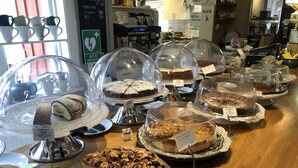 A bakery display with various cakes and pastries under glass domes, a coffee machine, and a menu board in the background.