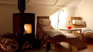A cozy room with a wood-burning stove, two wicker lounge chairs, a small table, and a basket of firewood.