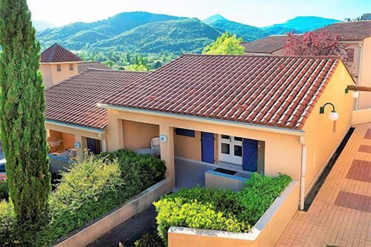A residential building with a tiled roof, surrounded by greenery and hills in the background.