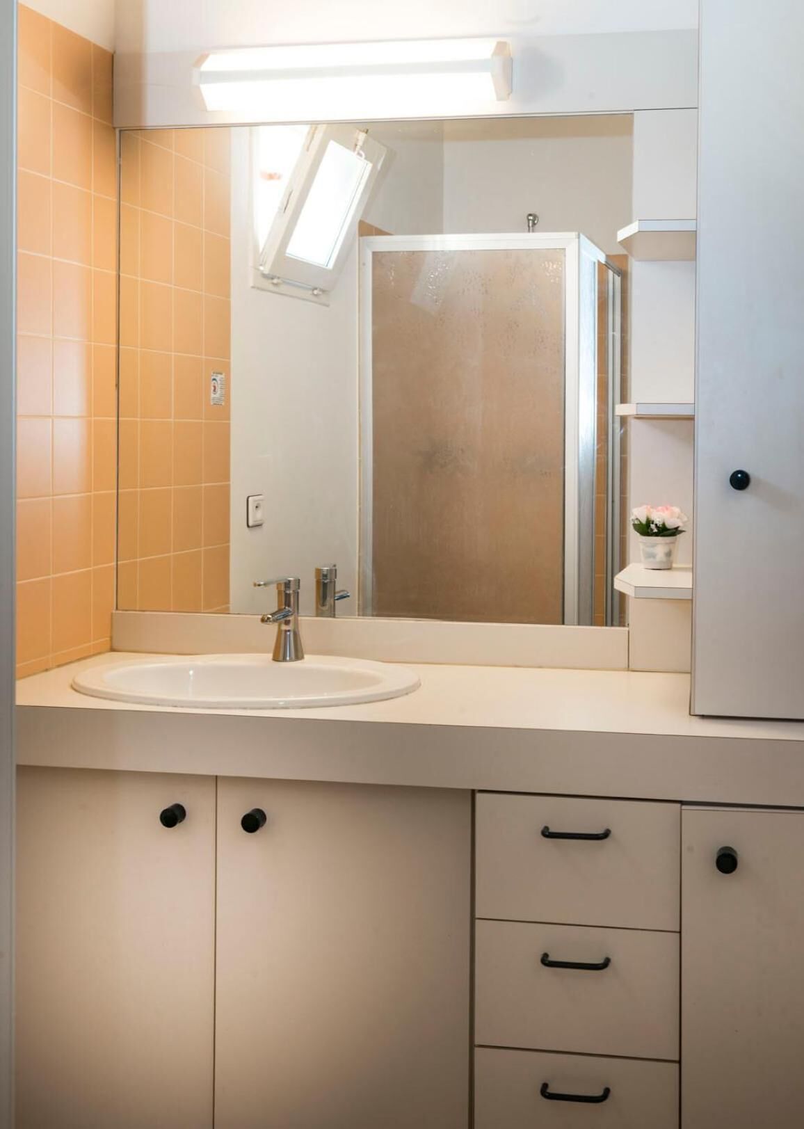 A bathroom with a white sink, a mirror, and a shower. There is a window above the mirror, and the walls are tiled in a light color.