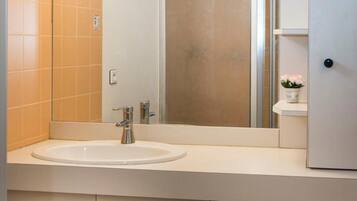 A bathroom with a white sink, a mirror, and a shower. There is a window above the mirror, and the walls are tiled in a light color.