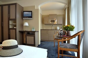 A hotel room with a bed, a desk with a lamp, a TV, a kitchenette, and a dining area with a chair and table.