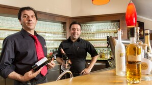 Twee personen aan een bar, met wijnflessen en glazen.