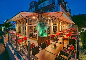 Ein Restaurant mit Außenbereich, Tischen, Stühlen und einem Baum in der Mitte, sowie ein Gebäude im Hintergrund.