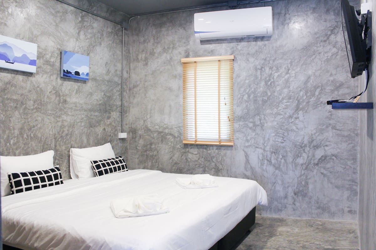 A hotel room with a marble wall, a bed with white bedding, two pillows, a window with blinds, and an air conditioning unit.