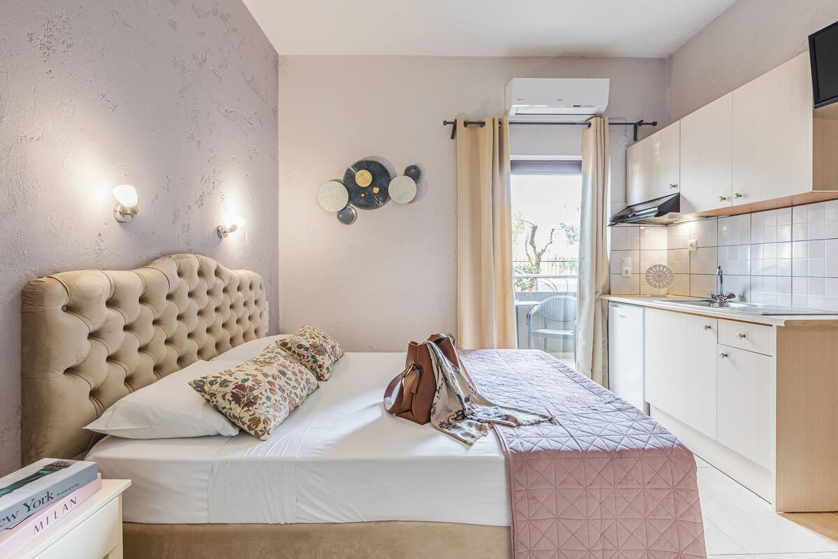 A bedroom with a tufted headboard, a bed with a patterned pillow, a bedside table with books, and a view of the kitchen.