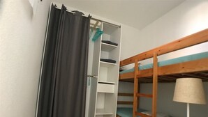 Een kleine, compacte kamer met een stapelbed, een garderobe met planken en een nachtkastje met een lamp.