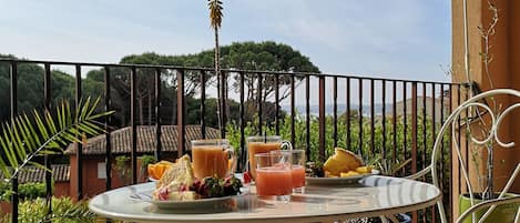 A terrace with a table set for breakfast, including drinks and fruit, surrounded by metal chairs and a railing.
