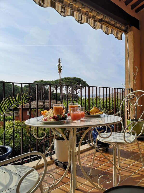 A terrace with a table set for breakfast, including drinks and fruit, surrounded by metal chairs and a railing.