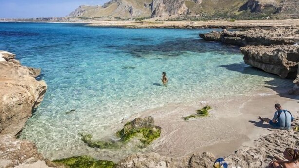 Una playa con agua cristalina, formaciones rocosas y montañas al fondo.