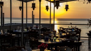 Un área de comedor al aire libre con mesas preparadas para una comida, con vistas al mar durante la puesta de sol.