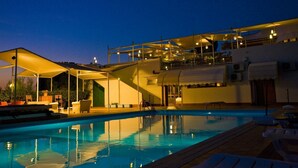Una piscina con zona techada y sillas de descanso, rodeada por un edificio de varios niveles e iluminación exterior.