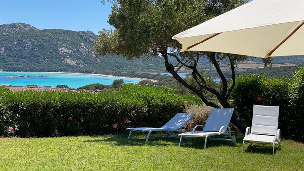 Un'area verde con due poltrone da relax e un ombrellone grande, con vista su un paesaggio costiero che include montagne e un mare blu limpido.