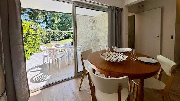Un coin repas avec une table ronde, des chaises blanches et une vue sur un jardin, visible à travers une porte vitrée.