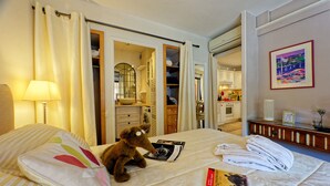 Ein Hotelzimmer mit einem Bett, einem kleinen Tisch mit Handtüchern, einer Lampe, einem Teddybären und Blick auf eine Küche und ein Badezimmer durch eine offene Tür.
