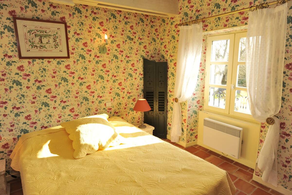 Ett sovrum med blommig tapet, en säng med ett gult överkast, en röd lampa och ett fönster med gardiner.