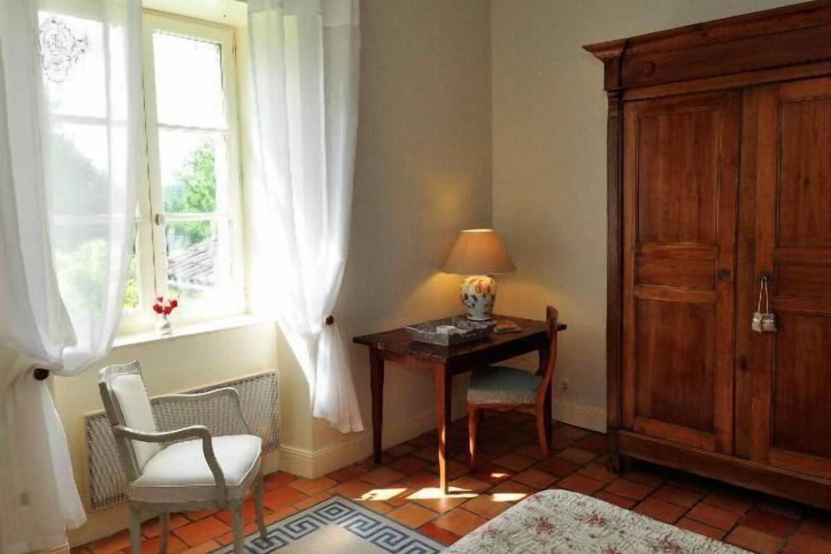 Ett rum med ett fönster, en stol, ett bord med en lampa och en garderob i trä.