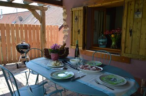 Un área de comedor al aire libre con una mesa preparada para dos personas, rodeada de sillas, una cerca de madera y una parrilla.