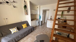 A compact living space with a grey sofa, a small round table, and a wooden ladder. - T2 + Mezzanine, 6 pers, Terrace, BBQ, Close to the beach (Port Barcarès)