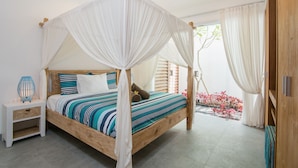 Uma cama com dossel e colunas, estrutura de madeira e cortinas brancas, uma mesa de cabeceira com abajur e vista para um jardim com flores vermelhas.