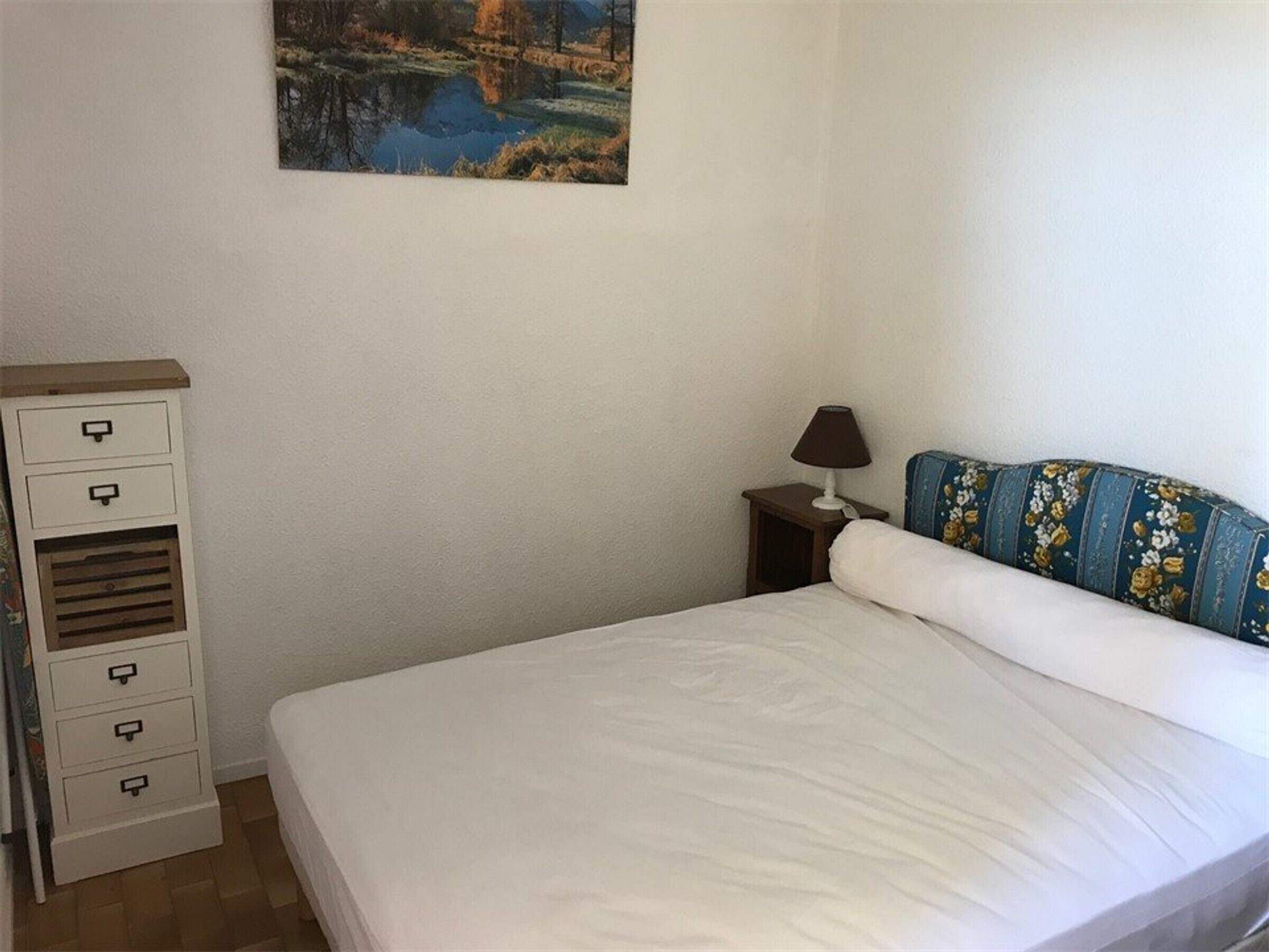 A bedroom with a bed, a dresser, a nightstand, a lamp, and a painting on the wall.