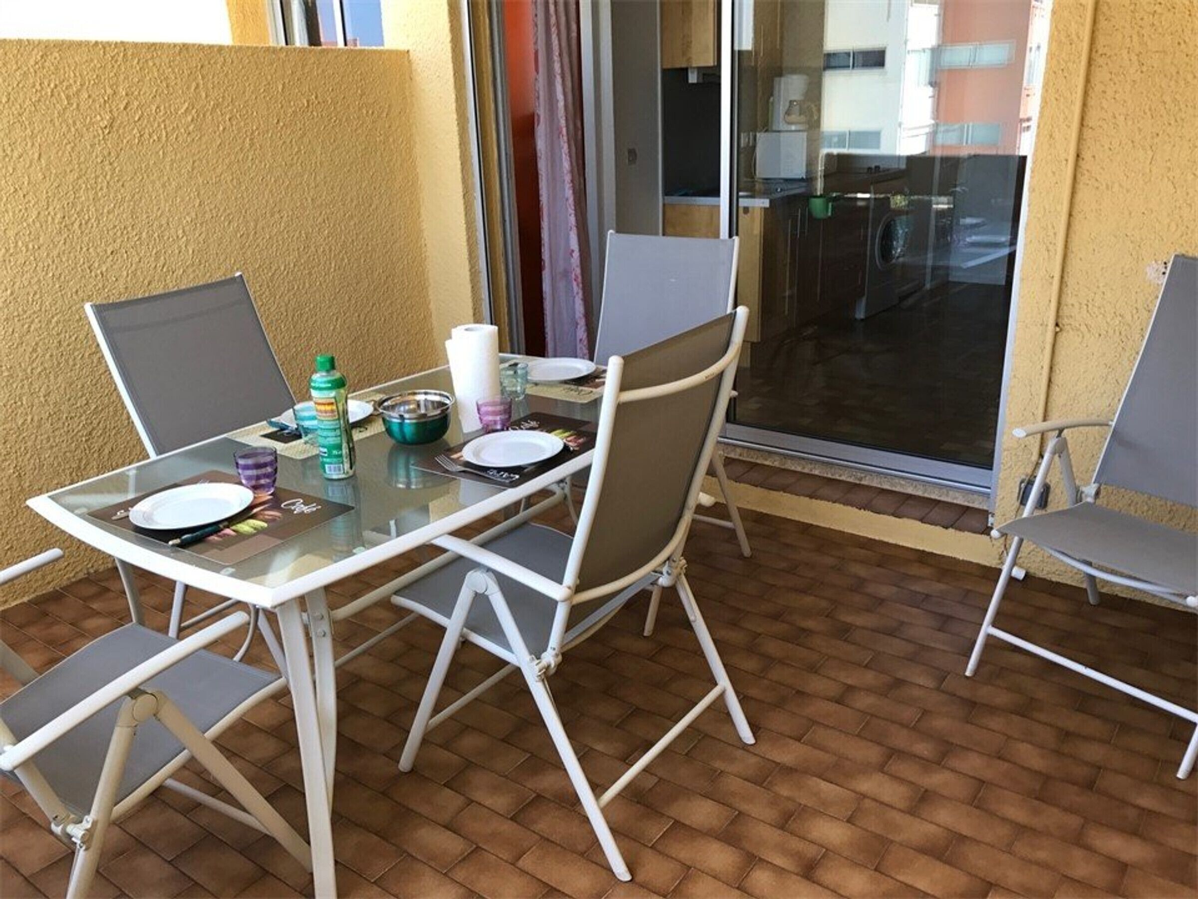 Een glazen tafel met vier stoelen, een fles, een kom en een bord op een balkon met een bakstenen vloer.