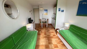 Ein Zimmer mit zwei grünen Betten, einem kleinen Schreibtisch mit Computer, einem Esstisch und einer Küchenzeile.