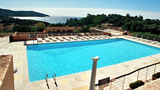 Ein rechteckiger Pool mit glasklarer, blauer Wasseroberfläche, umgeben von einer Steindeck und einem Metallgeländer. Im Hintergrund sind Gebäude und der Blick auf das Meer zu sehen.