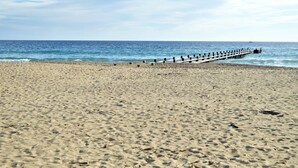 Ein Strand mit einer hölzernen Seebrücke, die sich ins Meer erstreckt, feinem Sand und blauem Himmel.