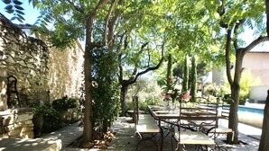 An outdoor patio with stone walls, wooden tables, and chairs, surrounded by trees and a pool. -  Mas Le Félibre **** (Caumont-sur-Durance)