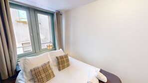 Ein ordentlich bezogenes Bett mit weißen Leinen und zwei karierten Kissen, eine Nachttischlampe und ein Fenster mit Vorhängen.