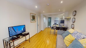 Ein Wohnzimmer mit Fernseher, Holzboden, einer Couch und einer Küche im Hintergrund.