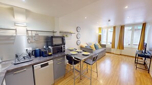 A compact kitchen with a dishwasher, a microwave, and a kettle. There is a dining area with a table and chairs, and a sofa with cushions.
